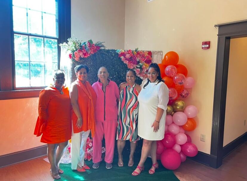 Five women in bright dresses posing beside a pink and orange balloon display